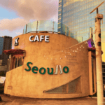 The exterior of K-Cafe on the Seoullo 7017 elevated walkway during sunset, with the golden light reflecting off a modern glass skyscraper in the background.