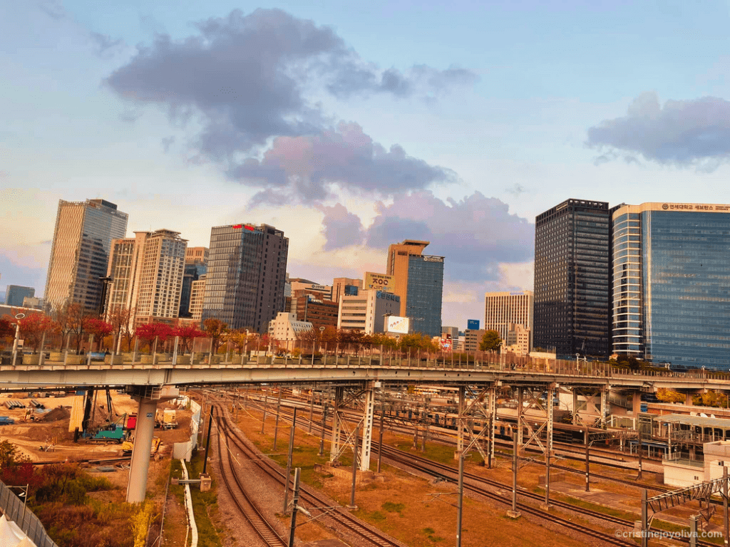 Seoul cityscape with high-rise buildings, railway tracks, and the Seoullo 7017 pedestrian walkway lined with autumn trees at sunset.