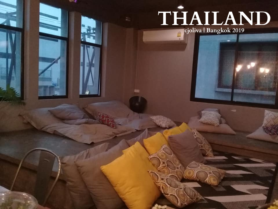 A cozy hostel common area with floor cushions, yellow pillows, and large windows at Silom Sp8ce in Bangkok.
