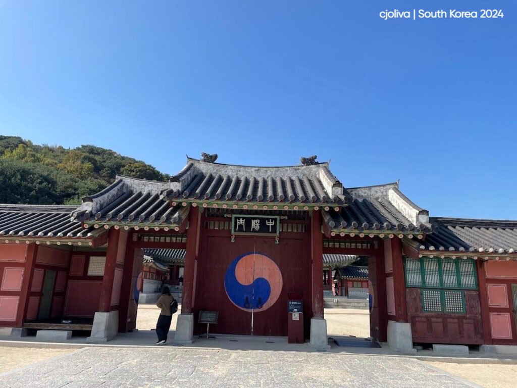Entrance gate of a traditional Korean palace with a curved tiled roof, a red wooden structure, the Taegeuk symbol painted on the doors, and a sign reading "Gapmun" under a clear blue sky.