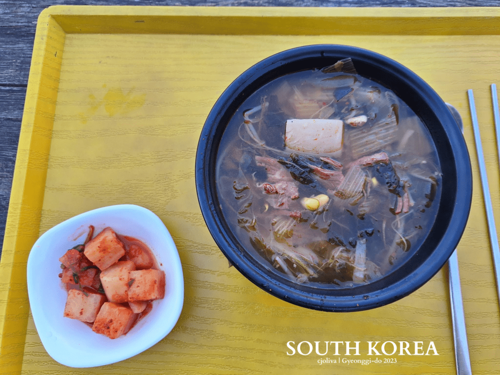 A top-down view of a steaming bowl of Korean beef and radish soup with bean sprouts and greens, served with a side of cubed radish kimchi (kkakdugi) on a yellow tray. Metal chopsticks and a spoon are visible.