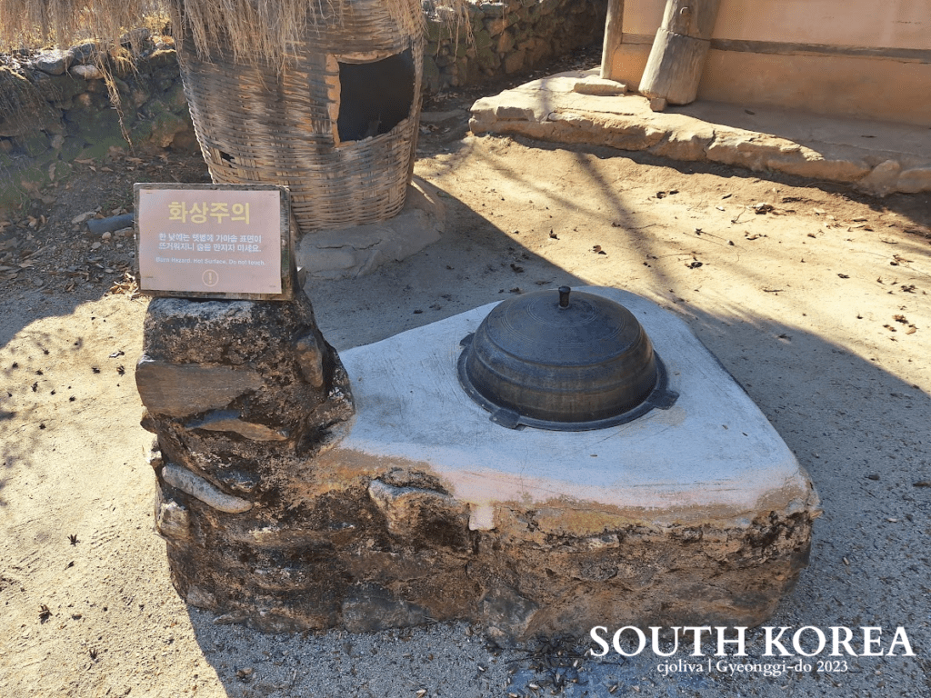 A close-up shot of a traditional Korean gamasot (large cast-iron cauldron) embedded in a stone and clay hearth outdoors. A warning sign in Korean and English sits next to it. In the background, rustic traditional structures and earth tones dominate.