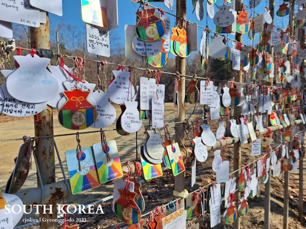 A wire fence covered in hundreds of colorful Korean wishing charms and small paper fortune bags (bokjumeoni). The charms have handwritten messages in Korean. The background shows a sunny outdoor field in winter.