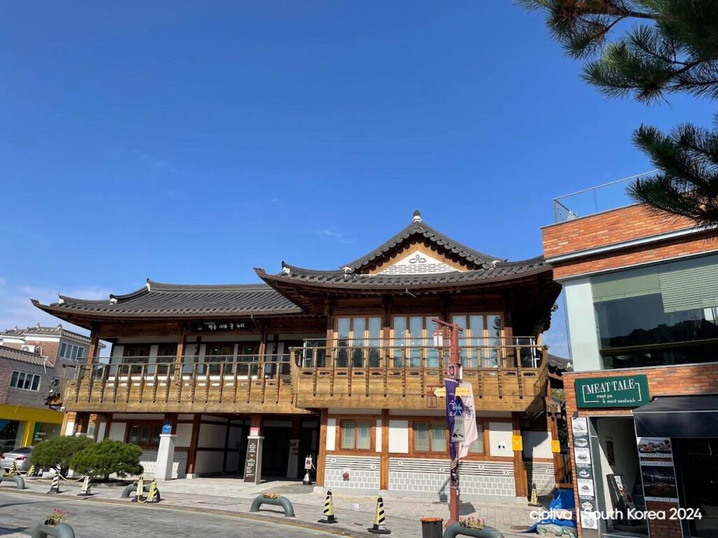 Traditional Korean hanok-style building with wooden balcony and tiled roof beside a modern brick and glass building on a sunny street in South Korea.