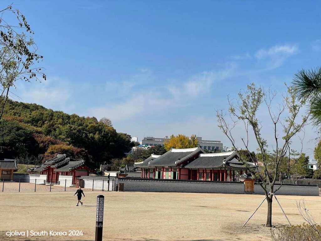 Traditional Korean palace complex with tiled roofs and ornate wooden detailing, surrounded by stone walls, autumn foliage, and modern buildings in the distance.