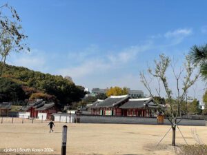 Traditional Korean palace complex with tiled roofs and ornate wooden detailing, surrounded by stone walls, autumn foliage, and modern buildings in the distance.
