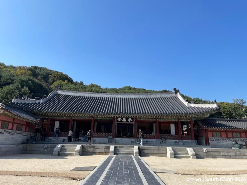Traditional Korean palace or temple with a tiled roof, red columns, and ornate eaves, set against forested hills under a clear blue sky with visitors in front.