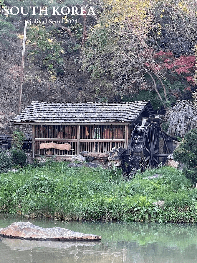 Rustic wooden watermill with large water wheel beside a pond in South Korea, surrounded by autumn foliage.