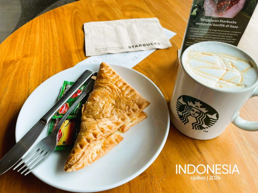 Starbucks table in Indonesia with a triangular puff pastry on a plate, fork and knife, condiment packets, and a white Starbucks mug topped with caramel drizzle.