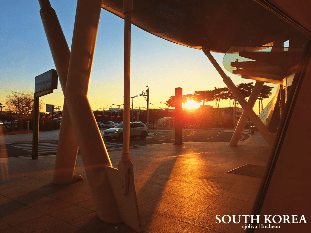 Golden sunrise at Incheon International Airport transit area with modern architectural pillars and a car in the background.
