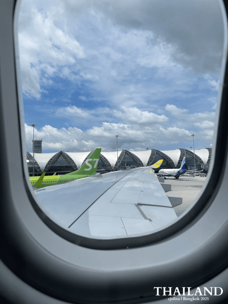 A view from an airplane window at Suvarnabhumi Airport in Bangkok, showing the distinct wavy terminal architecture, an airplane wing, and an S7 Airlines plane on the tarmac.
