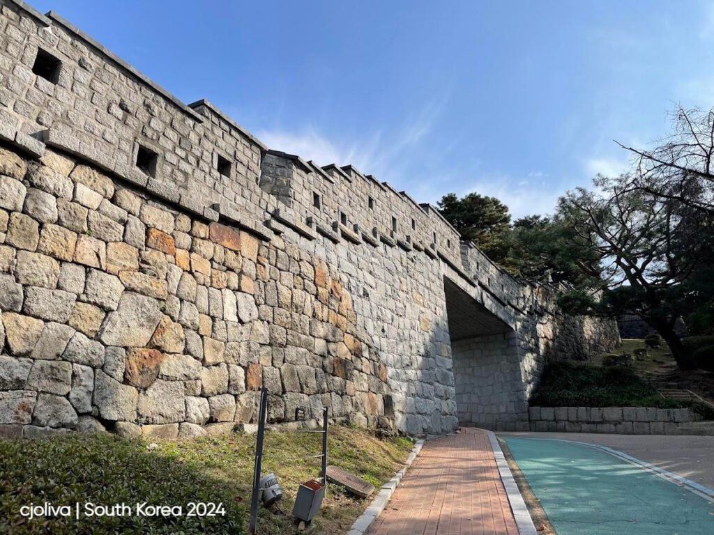 Historic stone wall archway at Hwaseong Fortress in Suwon, South Korea, with brick-paved pedestrian lane, green bicycle path, and surrounding trees under a clear blue sky.