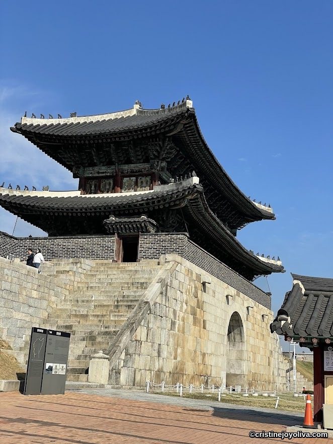 Traditional Korean fortress gate at Hwaseong Fortress in Suwon, South Korea, with stone base, arched entrance, and multi-tiered wooden pavilion featuring ornate eaves under a clear blue sky.