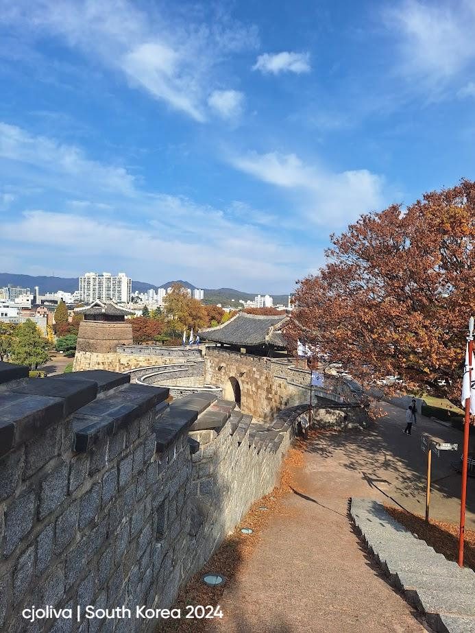 Curving stone wall of Hwaseong Fortress in Suwon, South Korea, with arched gateways, tiled rooftops, autumn-colored trees, and modern city buildings in the distance.