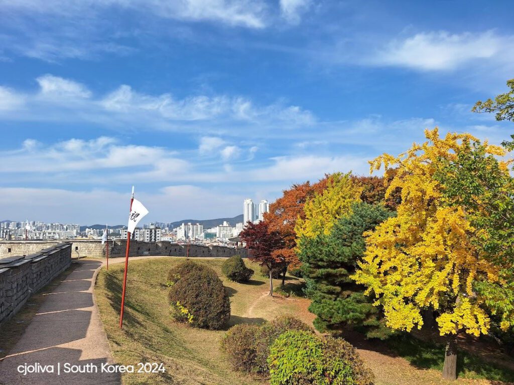 Stone wall pathway of Hwaseong Fortress in Suwon, South Korea, lined with flags bearing Korean characters, surrounded by vibrant autumn foliage and modern city buildings in the background.