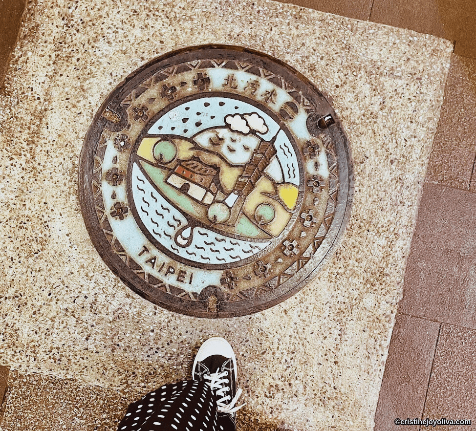 Colorful artistic manhole cover in Taipei featuring cartoon character, traditional building, and landscape motifs
