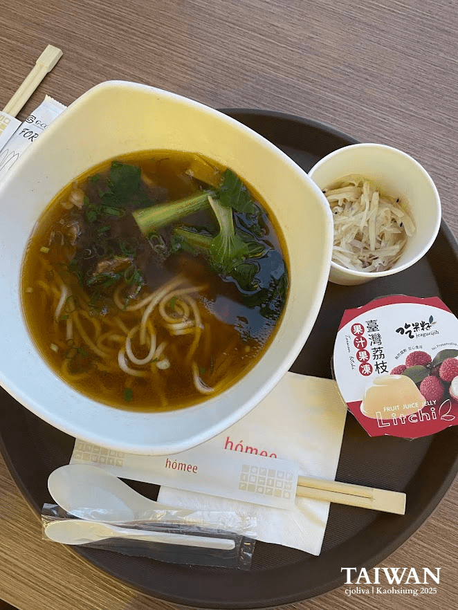 Taiwanese noodle soup with dark broth, leafy greens, scallions, side of shredded vegetables, and lychee fruit jelly drink on tray