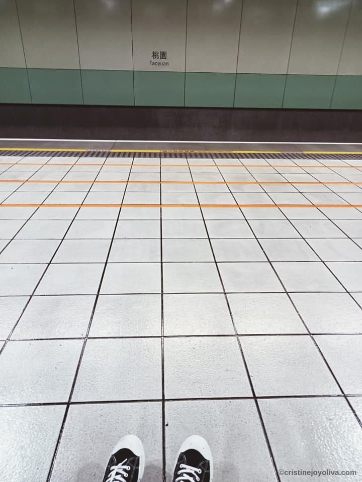A solo traveler's perspective of the tiled platform at Taoyuan Station in Taiwan, featuring black and white sneakers and the station sign.