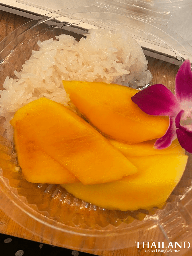 A plastic container of Thai mango sticky rice featuring sliced ripe yellow mango, glutinous rice, and a purple orchid garnish on a wooden table in Bangkok.