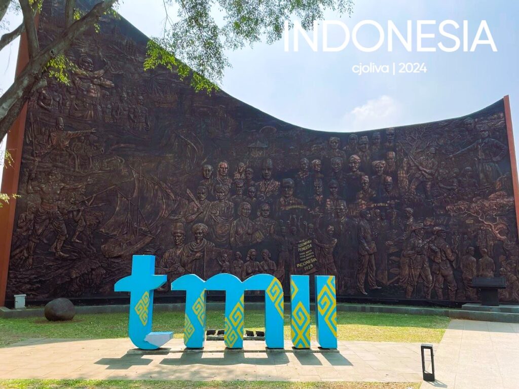 A detailed bronze relief sculpture on a wall depicting various historical and cultural scenes of Indonesia, with a blue circular sign featuring the white TMII (Taman Mini Indonesia Indah) logo in the foreground.