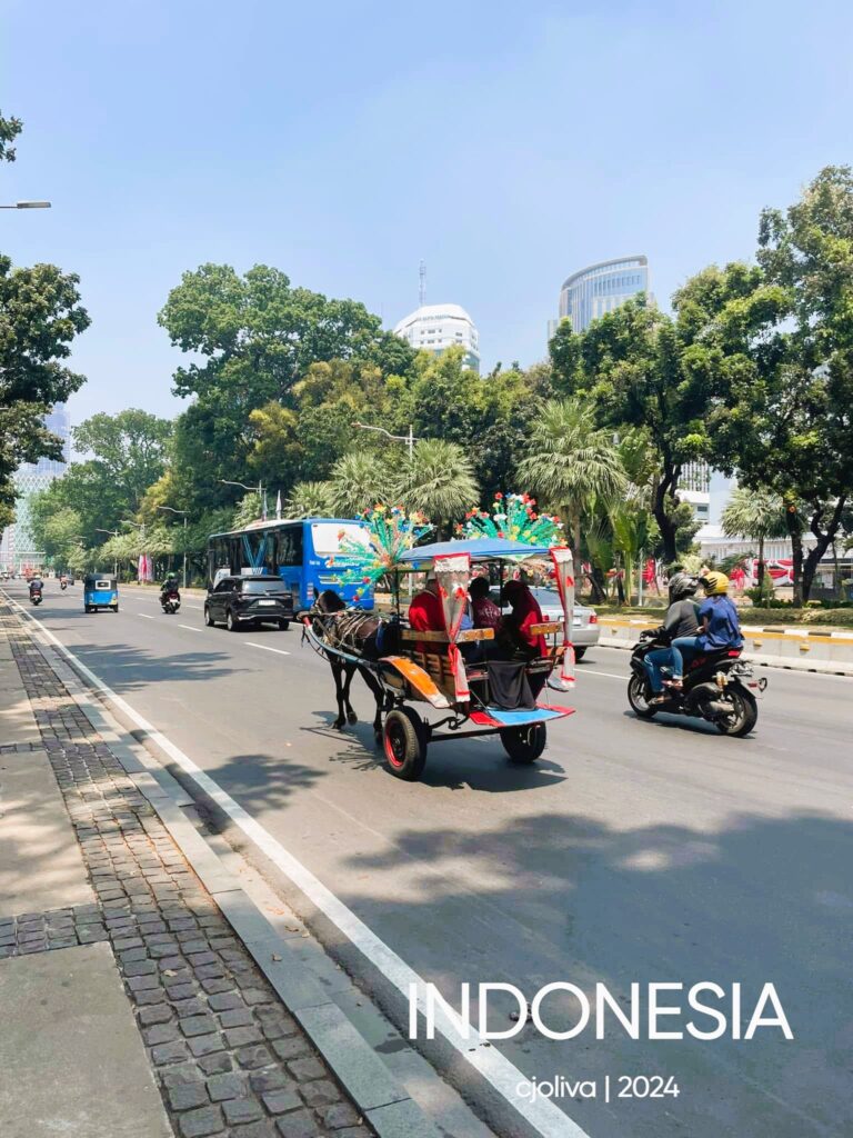 A colorful traditional Indonesian horse-drawn carriage (Delman) with passengers, driving on a wide paved city road in Jakarta surrounded by green trees, motorbikes, and a blue city bus.