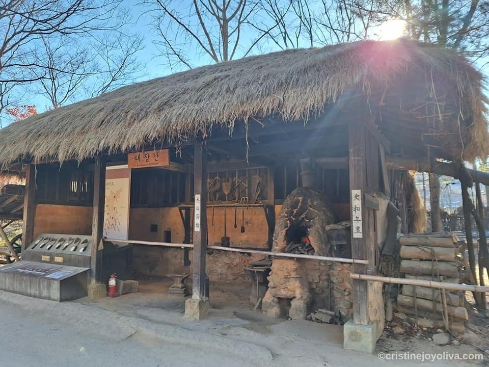 Traditional Korean blacksmith shop with thatched roof, stone furnace, and hanging tools