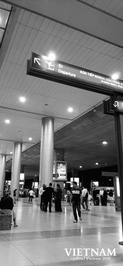 Black and white photo of Vietnam airport departure terminal showing people walking and standing under a well‑lit ceiling with large cylindrical columns and bilingual signage in Vietnamese and English directing passengers to departures, guest reception, and shops.