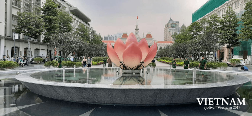 Public square in Vietnam featuring a large decorative fountain with a pink lotus sculpture, a landscaped pedestrian area with trees, people walking and relaxing, and background buildings with colonial and modern architecture, including a clock tower with a red flag.