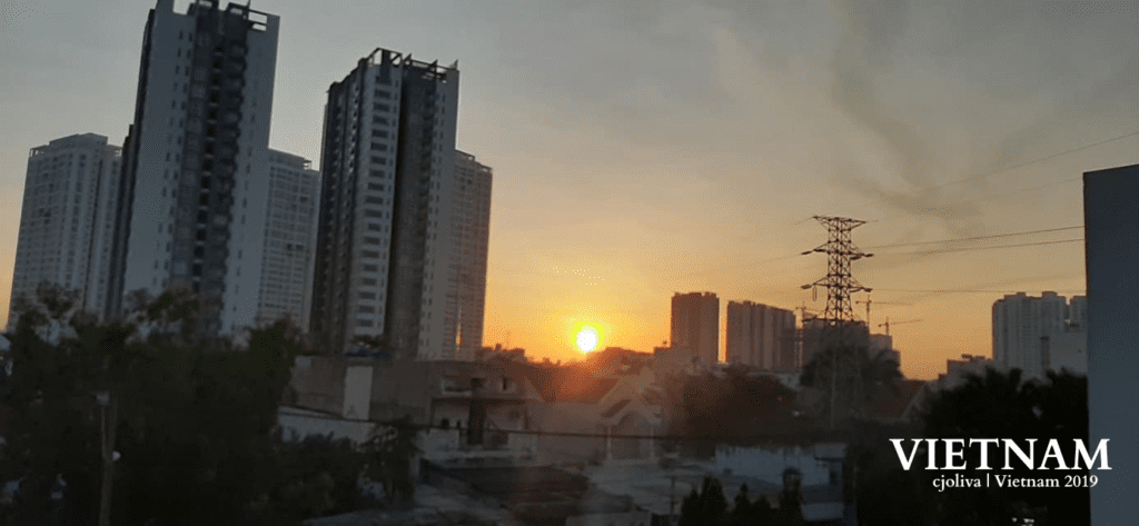Cityscape of Vietnam at sunrise, showing high-rise buildings, smaller foreground structures, and an electrical transmission tower with power lines under a warm orange sky.