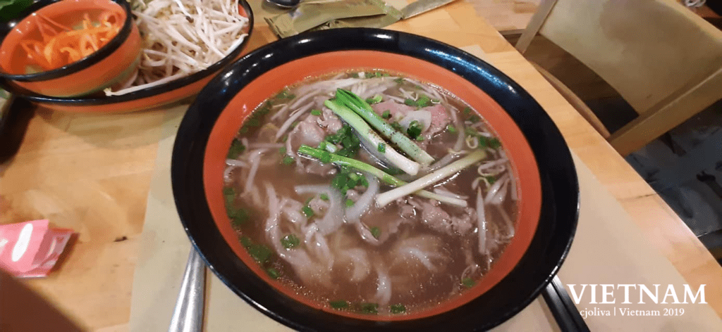 A bowl of Vietnamese pho noodle soup with broth, rice noodles, beef, green onions, and bean sprouts, surrounded by side dishes of bean sprouts and pickled vegetables on a wooden restaurant table.