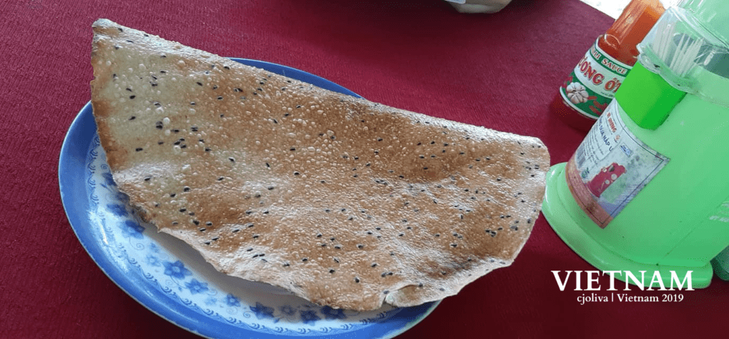 Large Vietnamese rice cracker with black sesame seeds on a blue and white floral plate, placed on a red tablecloth with chopsticks in a green container and a bottle of chili sauce nearby.