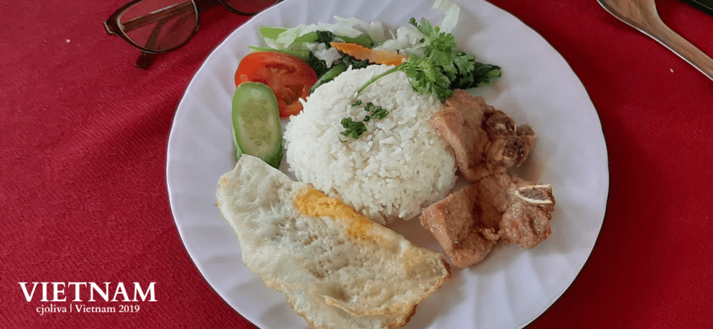 Plate of Vietnamese food with white rice topped with chopped green onions and cilantro, fried egg, grilled pork chops with bones, and fresh vegetables including cucumber, tomato, shredded cabbage, carrots, and leafy greens, served on a red tablecloth.