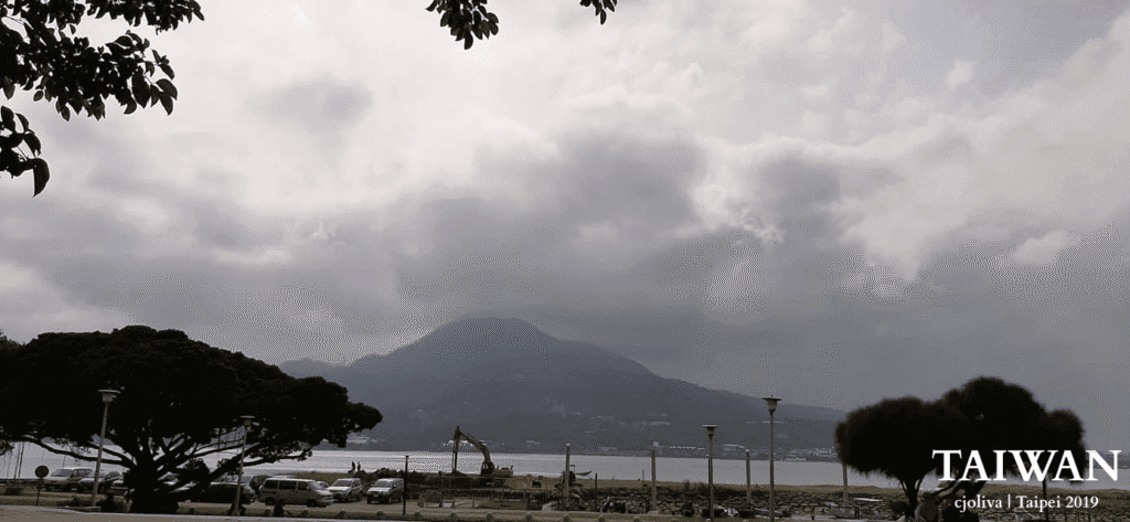 A wide-angle, desaturated view of the Tamsui River in Taipei, Taiwan, featuring Guanyin Mountain in the background under a heavy, cloudy sky. In the foreground, there are parked cars, a construction excavator, and leafy trees framing the riverbank.