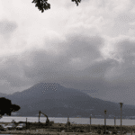 A wide-angle, desaturated view of the Tamsui River in Taipei, Taiwan, featuring Guanyin Mountain in the background under a heavy, cloudy sky. In the foreground, there are parked cars, a construction excavator, and leafy trees framing the riverbank.