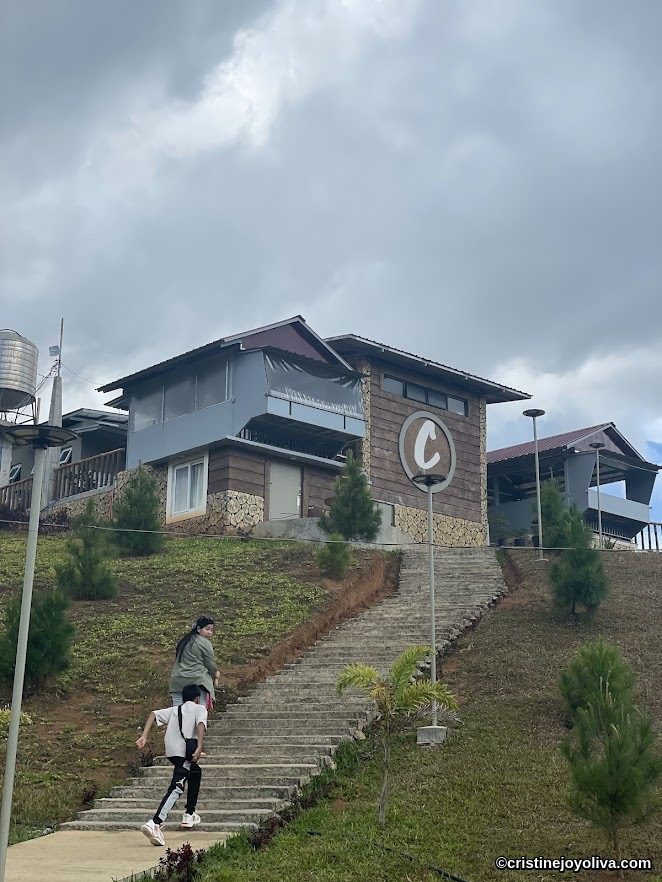 Two people are walking upstairs toward the C-Cafe mountain cabin, featuring a wood and stone exterior, surrounded by grassy slopes and cloudy skies.