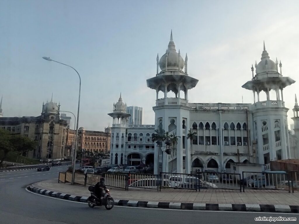 Kuala Lumpur Railway Station with white Mughal-style domes, ornate arches, and a motorcyclist riding along the curved road in front.