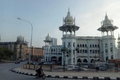 Kuala Lumpur Railway Station with white Mughal-style domes, ornate arches, and a motorcyclist riding along the curved road in front.