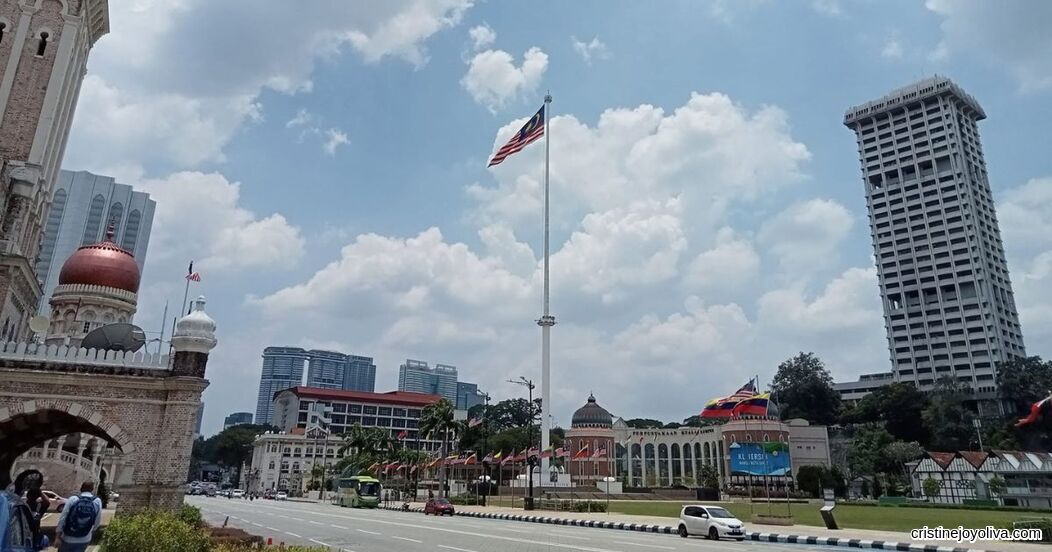 Merdeka Square in Kuala Lumpur, Malaysia, with the tall flagpole flying the national flag, the Sultan Abdul Samad Building, and surrounding colonial and modern architecture.