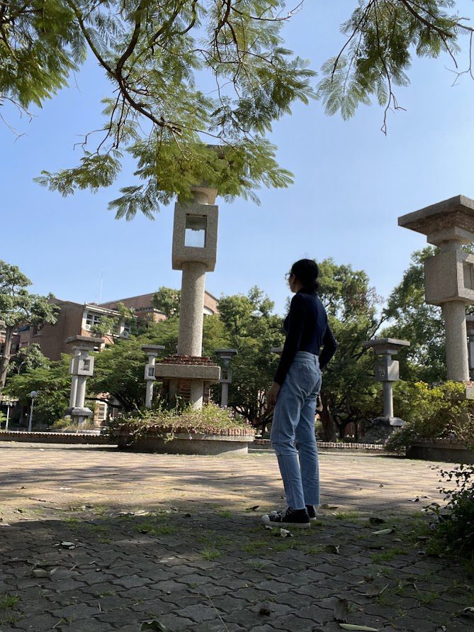 Person standing among tall stone lantern-like structures under green trees at National Chung Cheng University in Chiayi, Taiwan, a filming location for Meteor Garden.