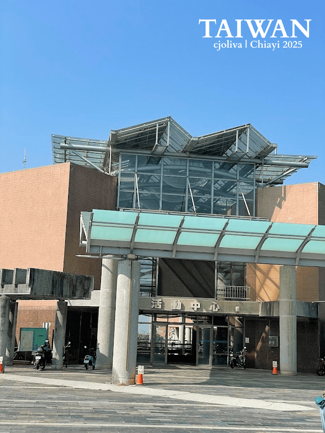 The Modern Activity Center at National Chung Cheng University in Chiayi, Taiwan, with a glass canopy, brick exterior, and scooters parked outside under a clear blue sky.