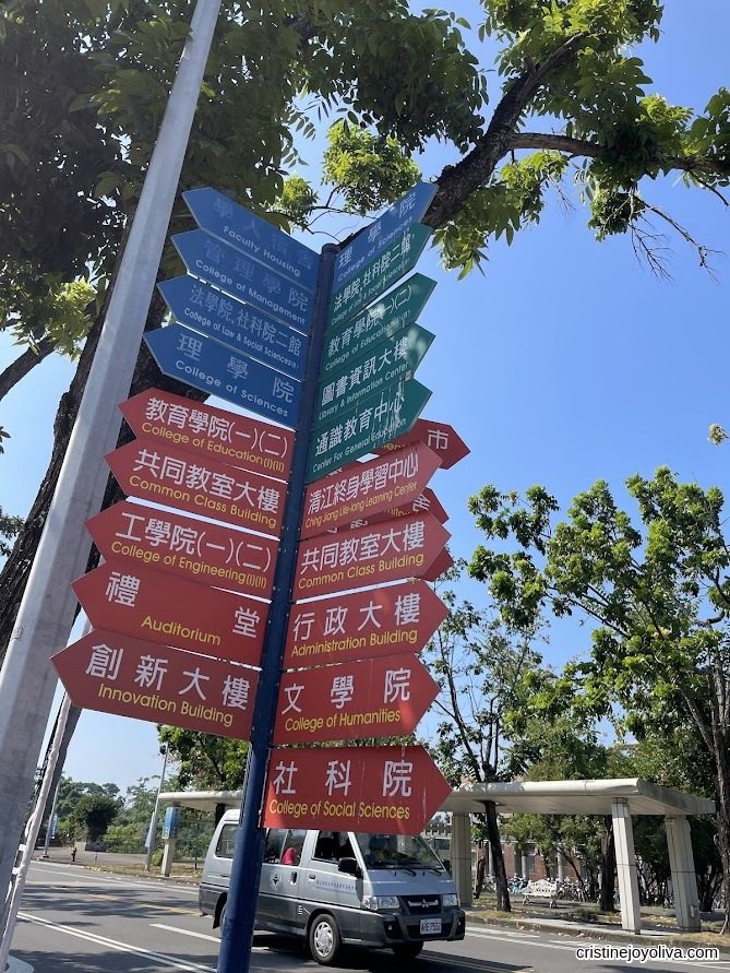 Directional signpost at National Chung Cheng University in Chiayi, Taiwan, with color-coded arrows pointing to various colleges and facilities, surrounded by trees and a clear blue sky.