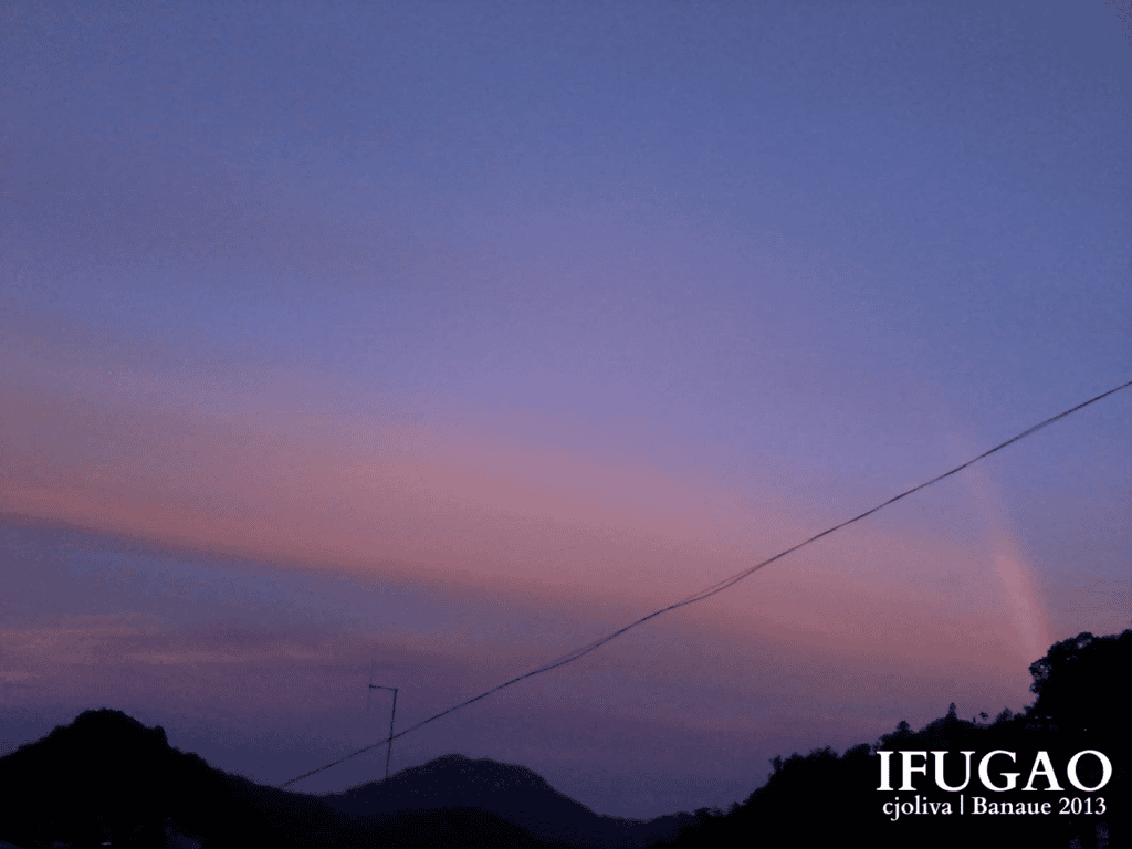 Soft purple and pink twilight sky over dark mountain silhouettes with a faint rainbow on the horizon in Banaue.