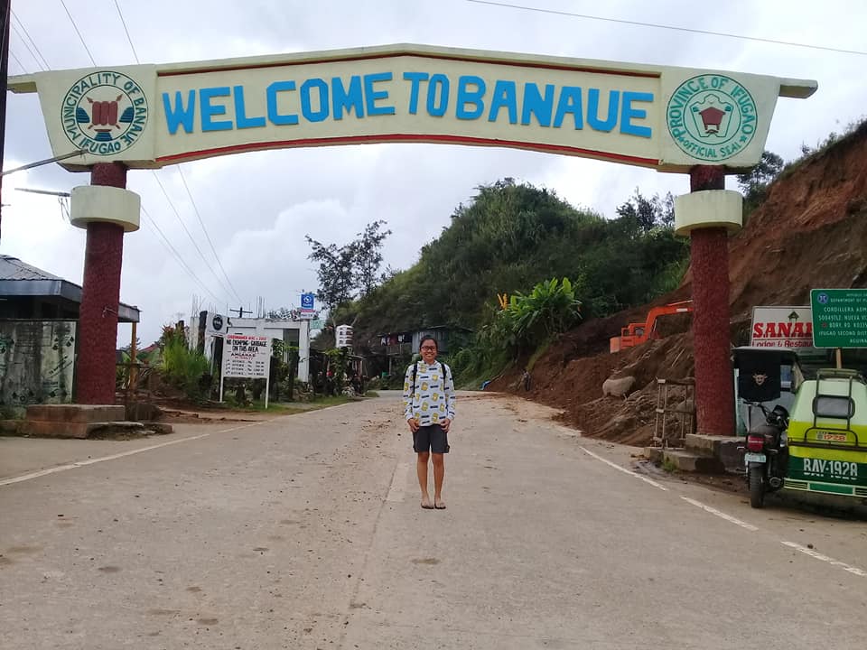 A person standing beneath the Banaue welcome arch supported by red pillars with official seals of the Municipality of Banaue and Province of Ifugao, hillside vegetation, a tricycle, and town signs are in the background.