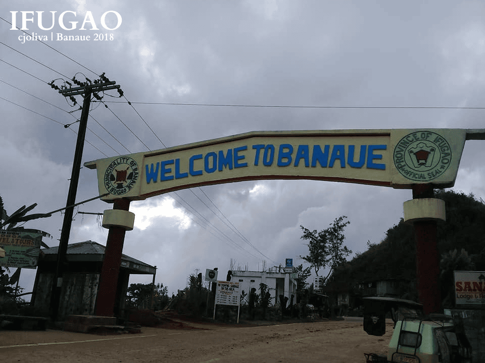 A large archway sign reading “Welcome to Banaue” with seals of the Municipality of Banaue and Province of Ifugao stands over a road leading into town under cloudy skies.