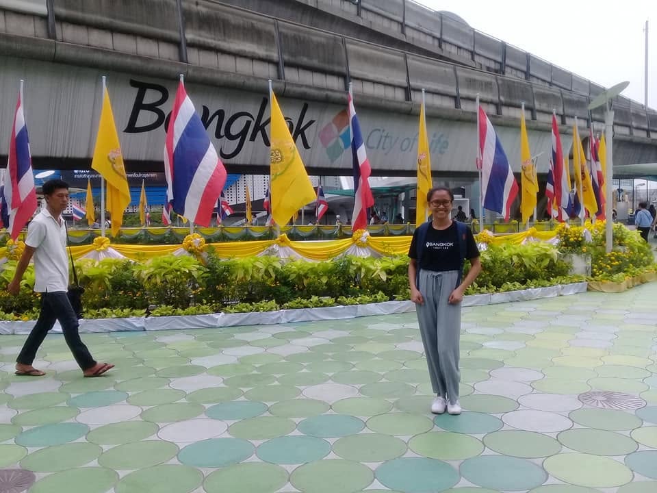 A woman smiling and posing near Thai and yellow flags with Bangkok city sign and pedestrian walkway in background