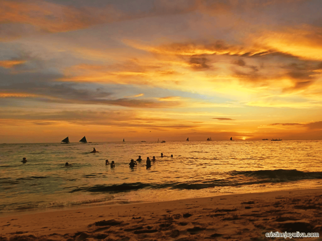 Boracay Beach at sunset with a vibrant orange and gold sky, sailboats with blue sails on the horizon, and people swimming near the shore under glowing light.