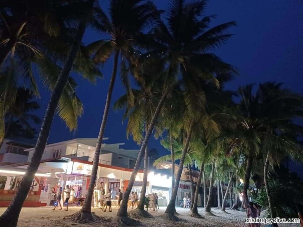 Boracay Beach at night with tall palm trees illuminated by lights, lively restaurants and shops with bright signage, and people walking on sandy ground under a dark blue sky.