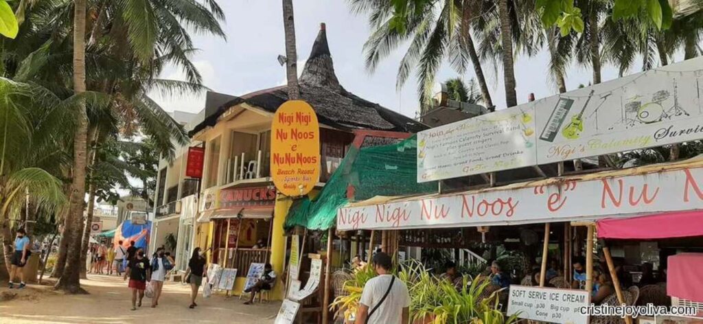 Boracay tropical street scene with palm trees, a sandy pathway, and Nigi Nigi Nu Noos Beach Resort featuring a thatched roof, a yellow vertical sign, colorful banners, and visitors enjoying the lively atmosphere.