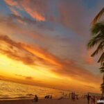 Boracay Beach at sunset with a vivid orange, pink, and blue sky; sailboats on the horizon; palm trees on the side; and people enjoying the sandy shore.