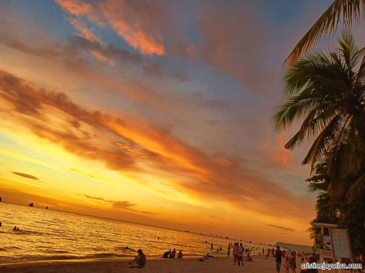 Boracay Beach at sunset with a vivid orange, pink, and blue sky; sailboats on the horizon; palm trees on the side; and people enjoying the sandy shore.